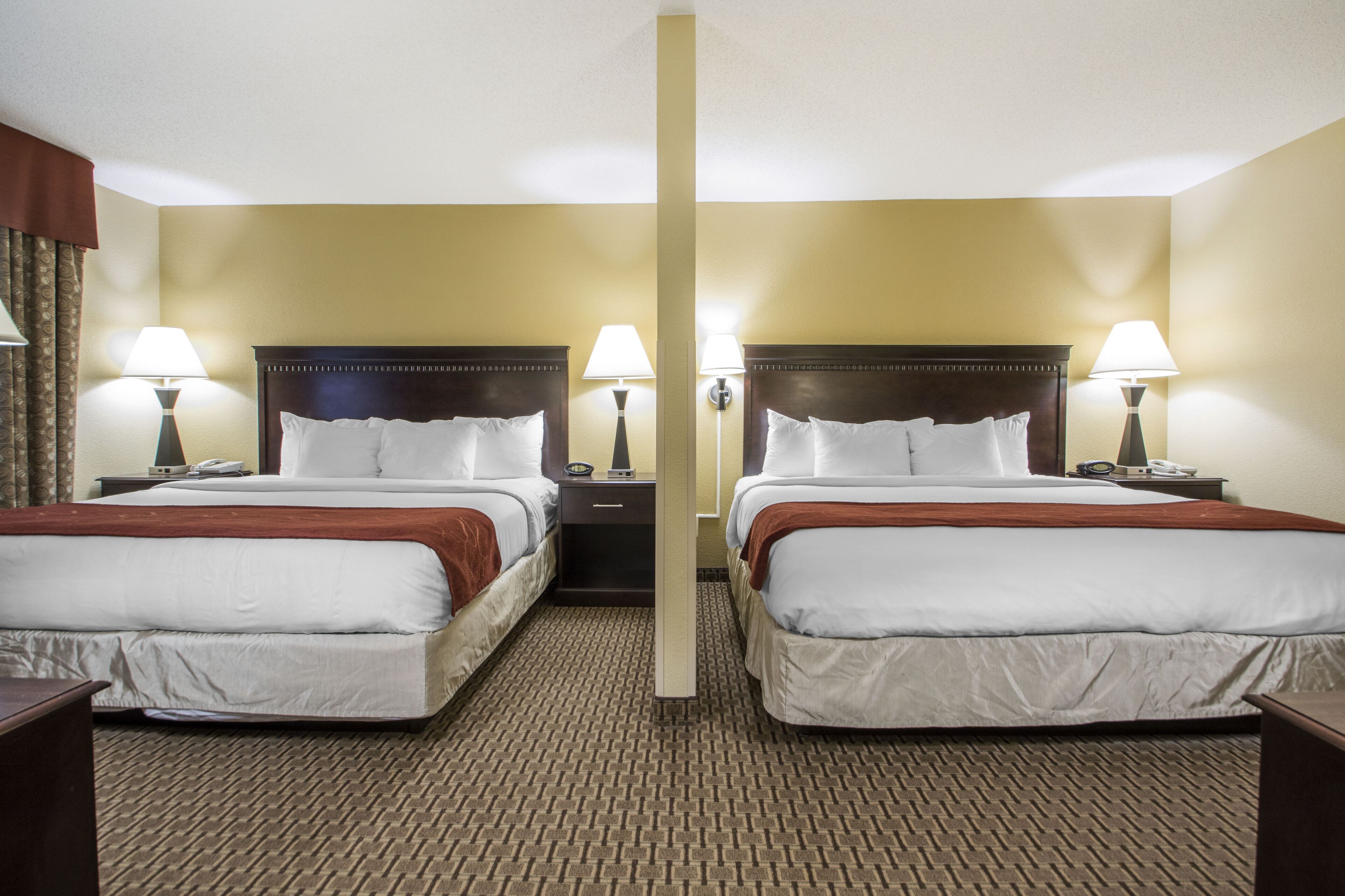 A hotel room with two beds, each with a wooden headboard, white bedding, and a brown throw blanket. There are bedside lamps, a nightstand with a phone, and a carpeted floor.