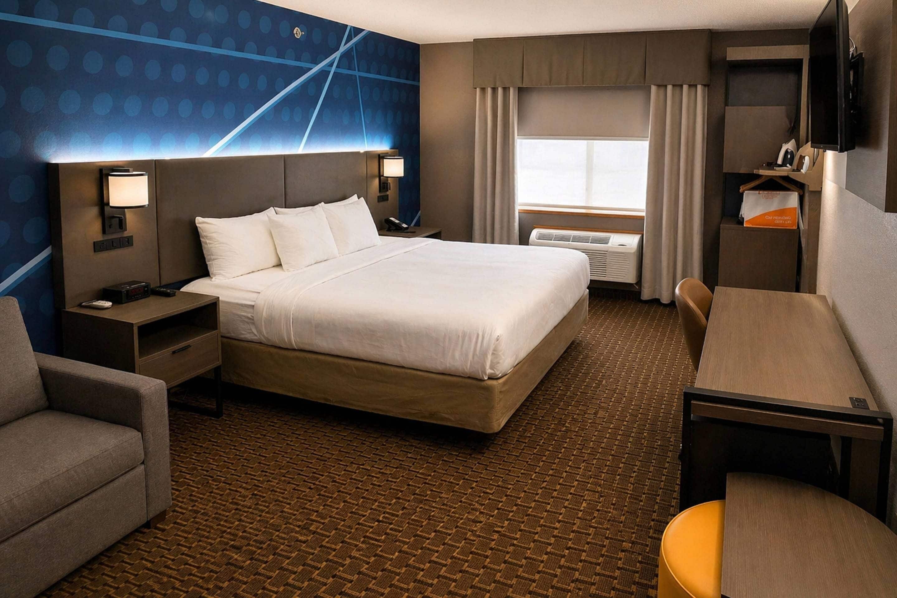 A hotel room with a large bed, a desk, a chair, a TV, a desk lamp, a nightstand, and a wall-mounted shelf.