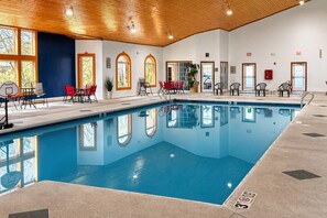Una piscina interior con agua cristalina, rodeada de sillas y mesas, y grandes ventanales que dejan entrar luz natural.
