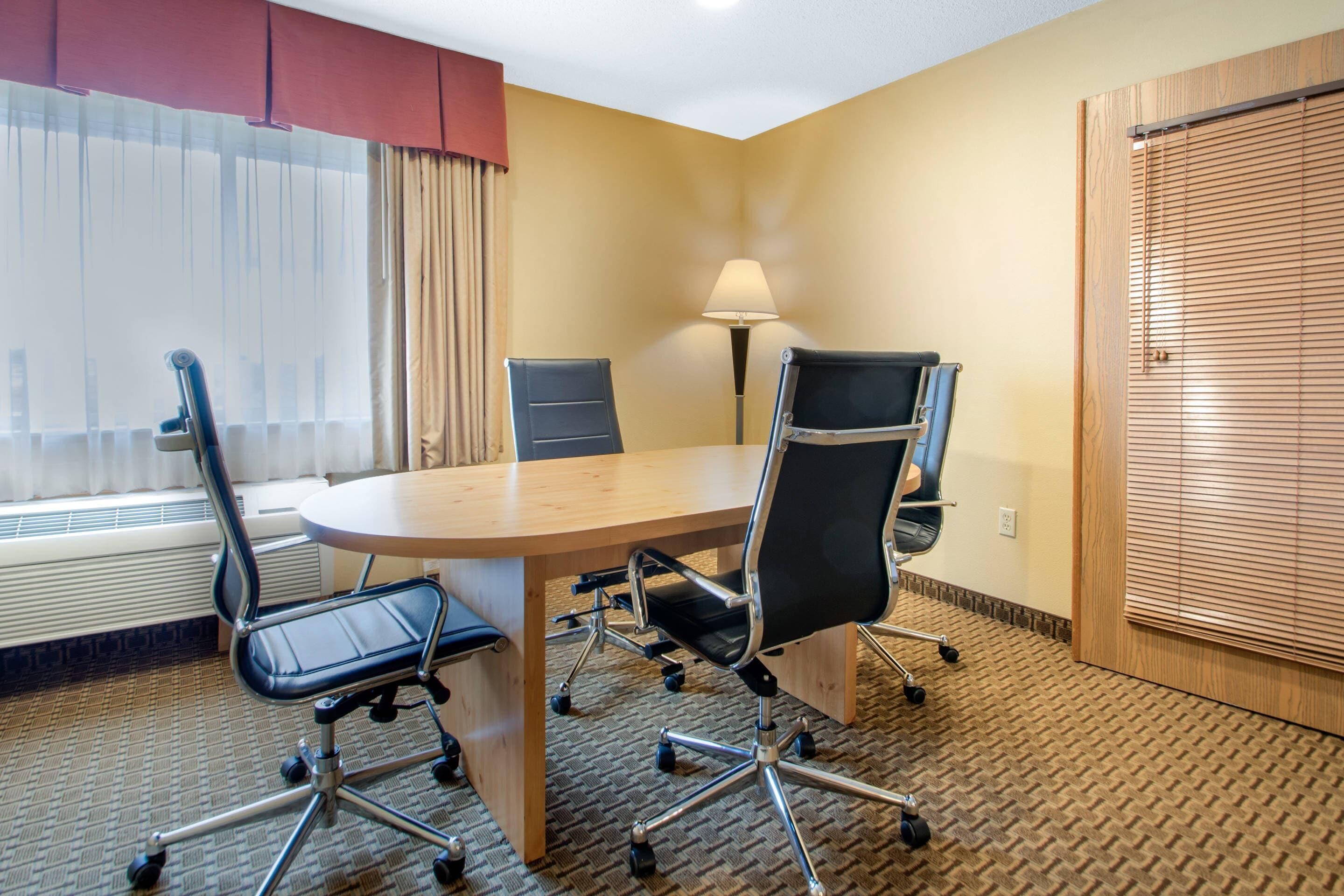 A hotel conference room with a wooden table, four black office chairs, a lamp, and a window with curtains.