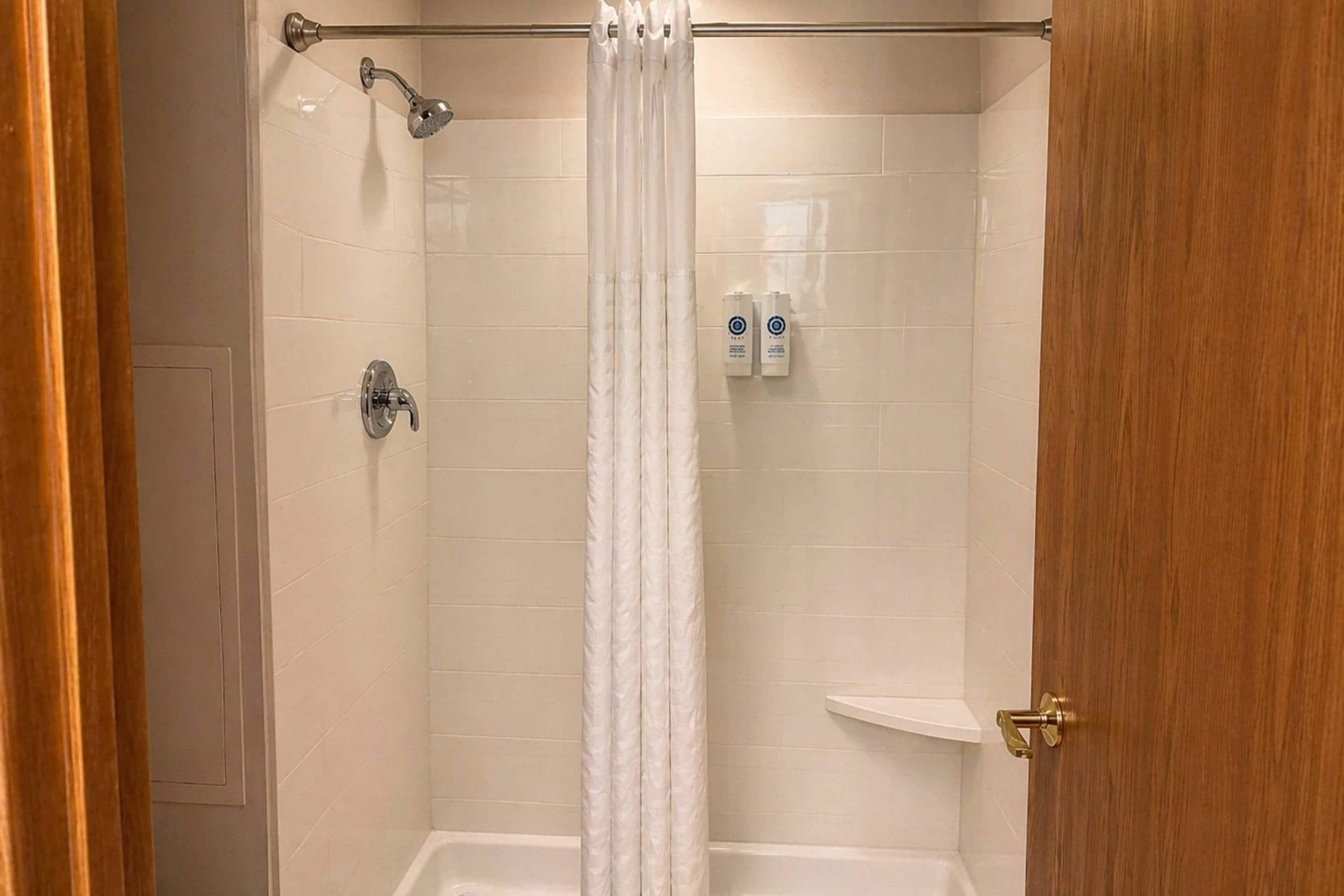 A shower with a white curtain, a handheld showerhead, and two bottles of shampoo on the wall.