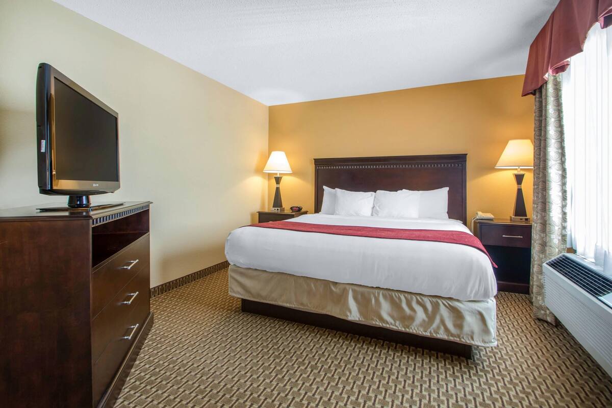 A hotel room with a bed, two bedside tables with lamps, a flat-screen TV, a dresser, and a window with curtains.