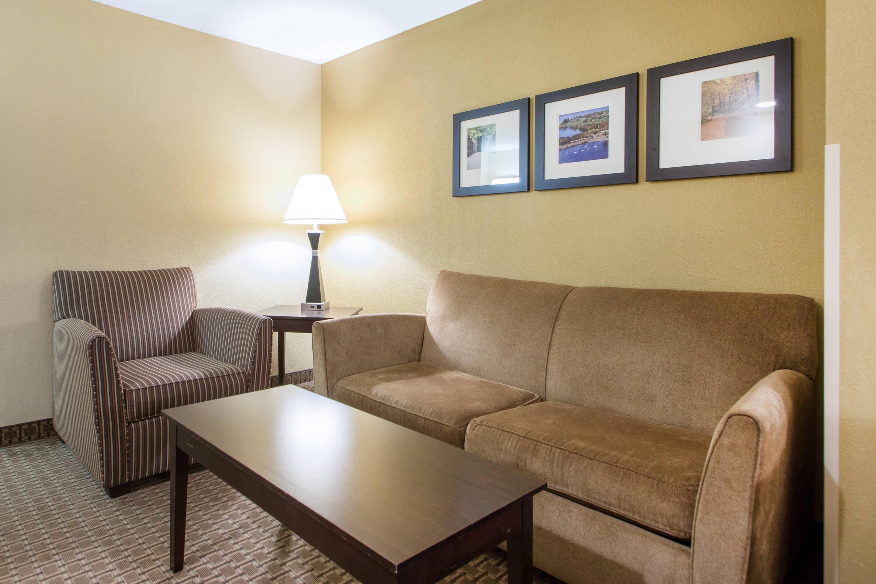 Una habitación de hotel con un sillón a rayas, un sofá marrón, una mesa de centro de madera y una lámpara. Hay tres cuadros en la pared.