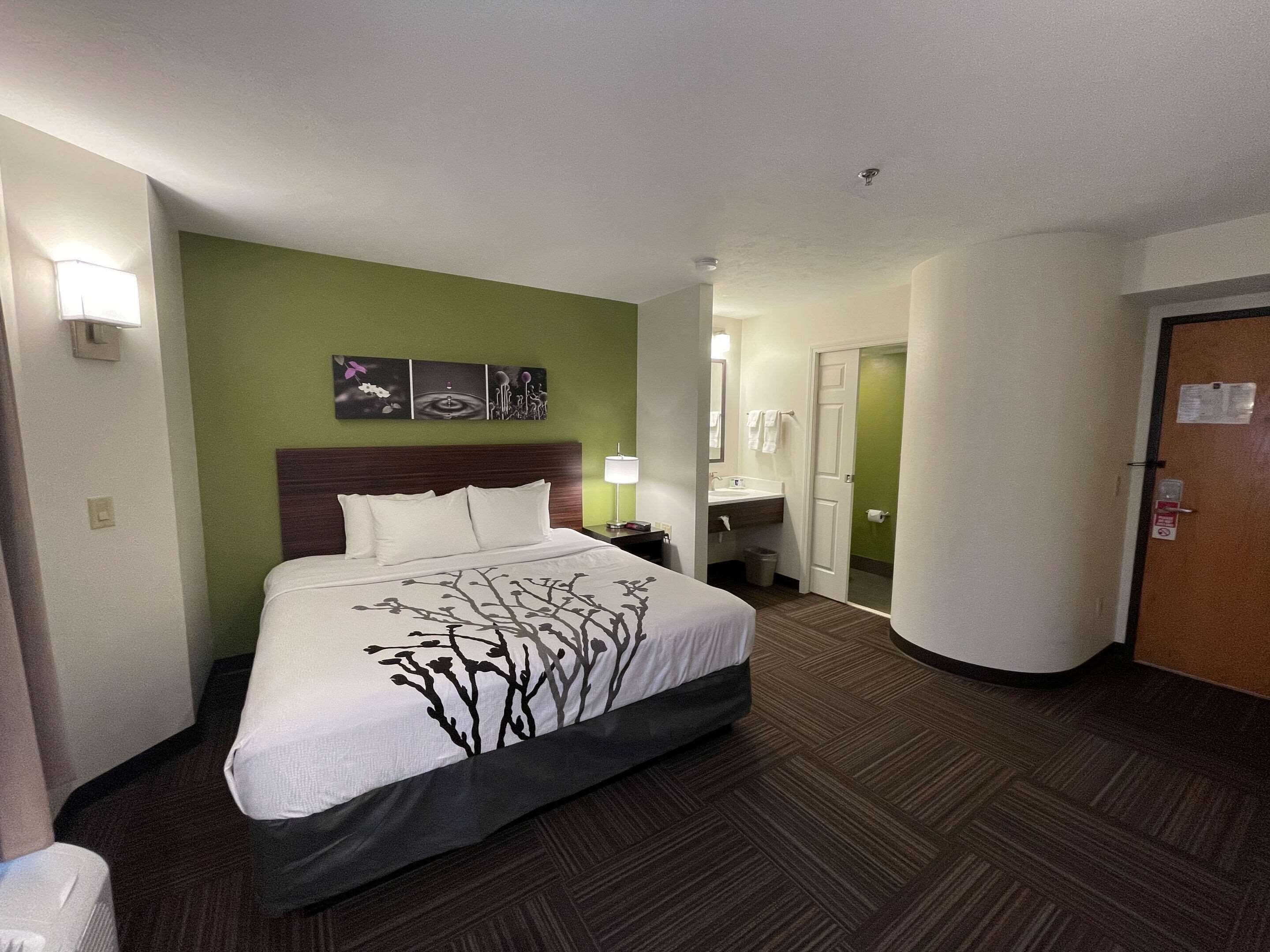 Ein Hotelzimmer mit einem großen Bett, einem Nachttisch, einer Wandlampe, einem Badezimmer mit Waschbecken und Spiegel sowie einer Tür mit einem Schild.