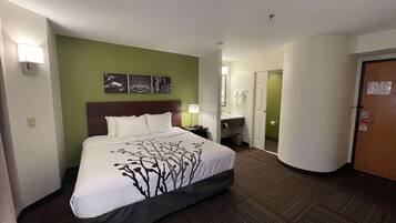 Quarto de hotel com uma cama grande, uma mesa de cabeceira, uma luminária na parede, um banheiro com pia e espelho, e uma porta com um aviso.