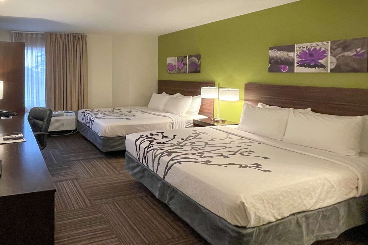 A hotel room with two beds, a desk, and a chair. There are wall art pieces and a window with curtains.