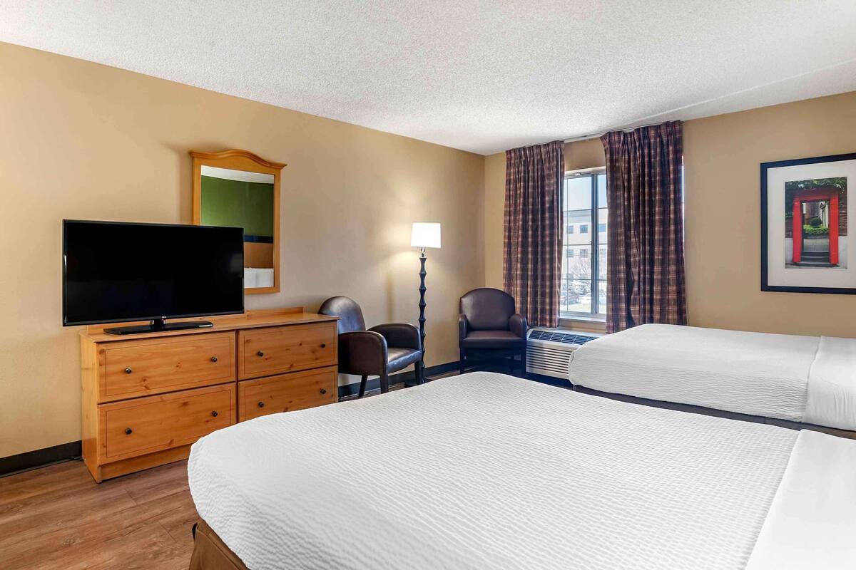 A hotel room with two beds, a TV on a wooden dresser, a chair, a lamp, and a window with curtains.