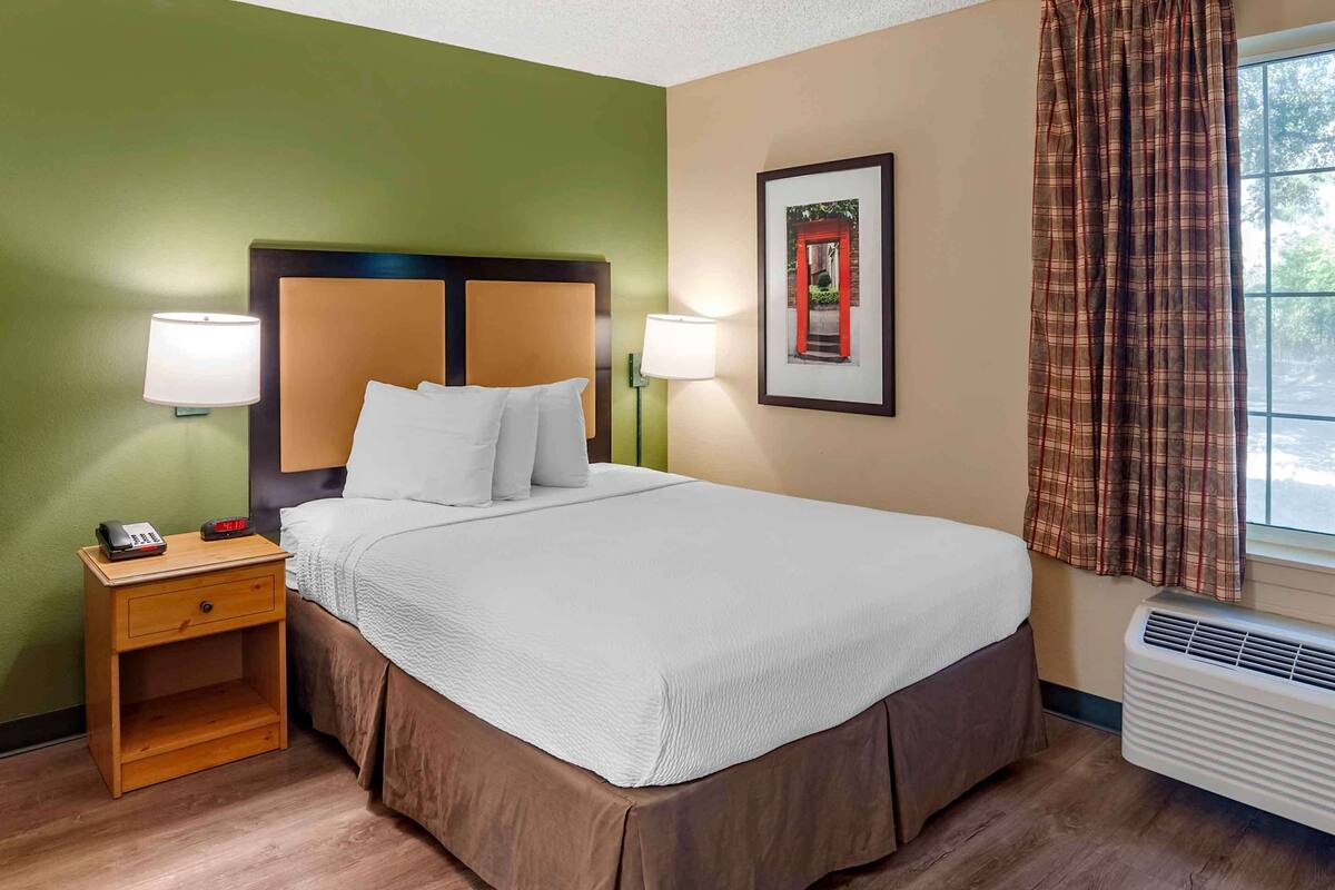 A hotel room with a bed, nightstand, wall-mounted lamps, a framed picture, and a window with curtains.
