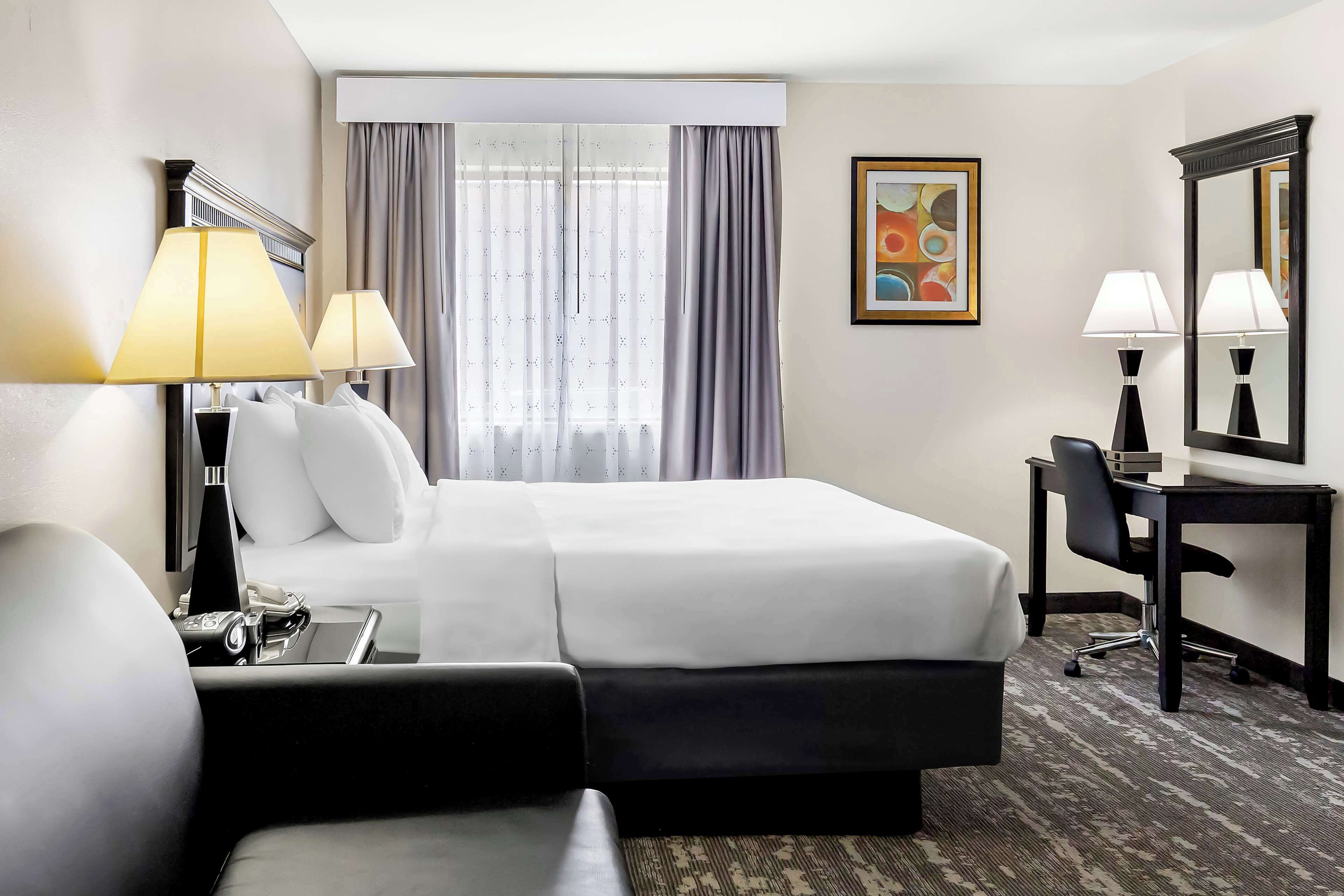 Habitación de hotel con cama, dos lámparas de noche, escritorio con silla, un espejo grande y ventana con cortinas.