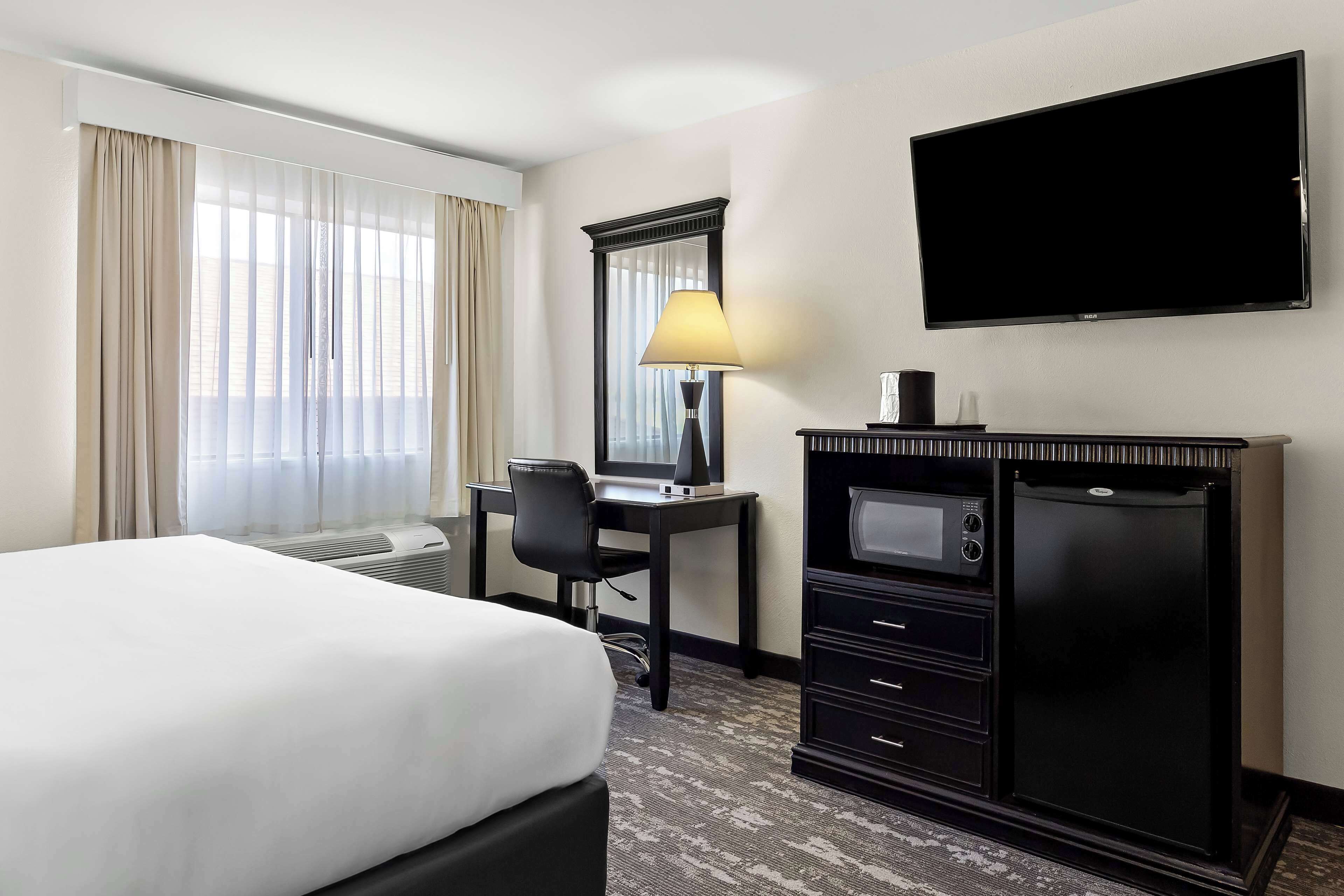 A hotel room with a bed, a desk with a computer, a flat-screen TV, a minibar, and a large window with curtains.