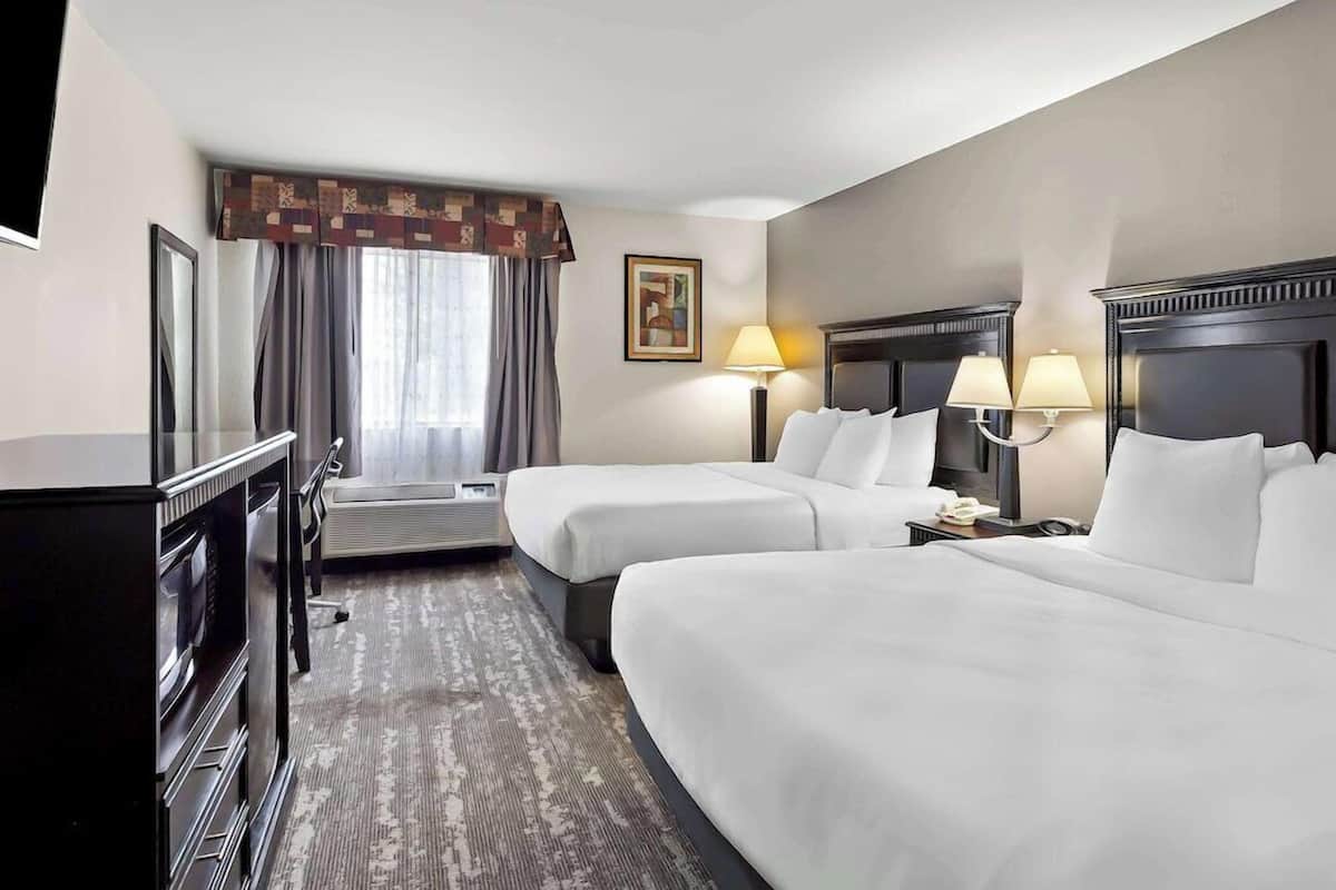 A hotel room with two beds, a desk, a chair, a TV, a lamp, and a painting on the wall.