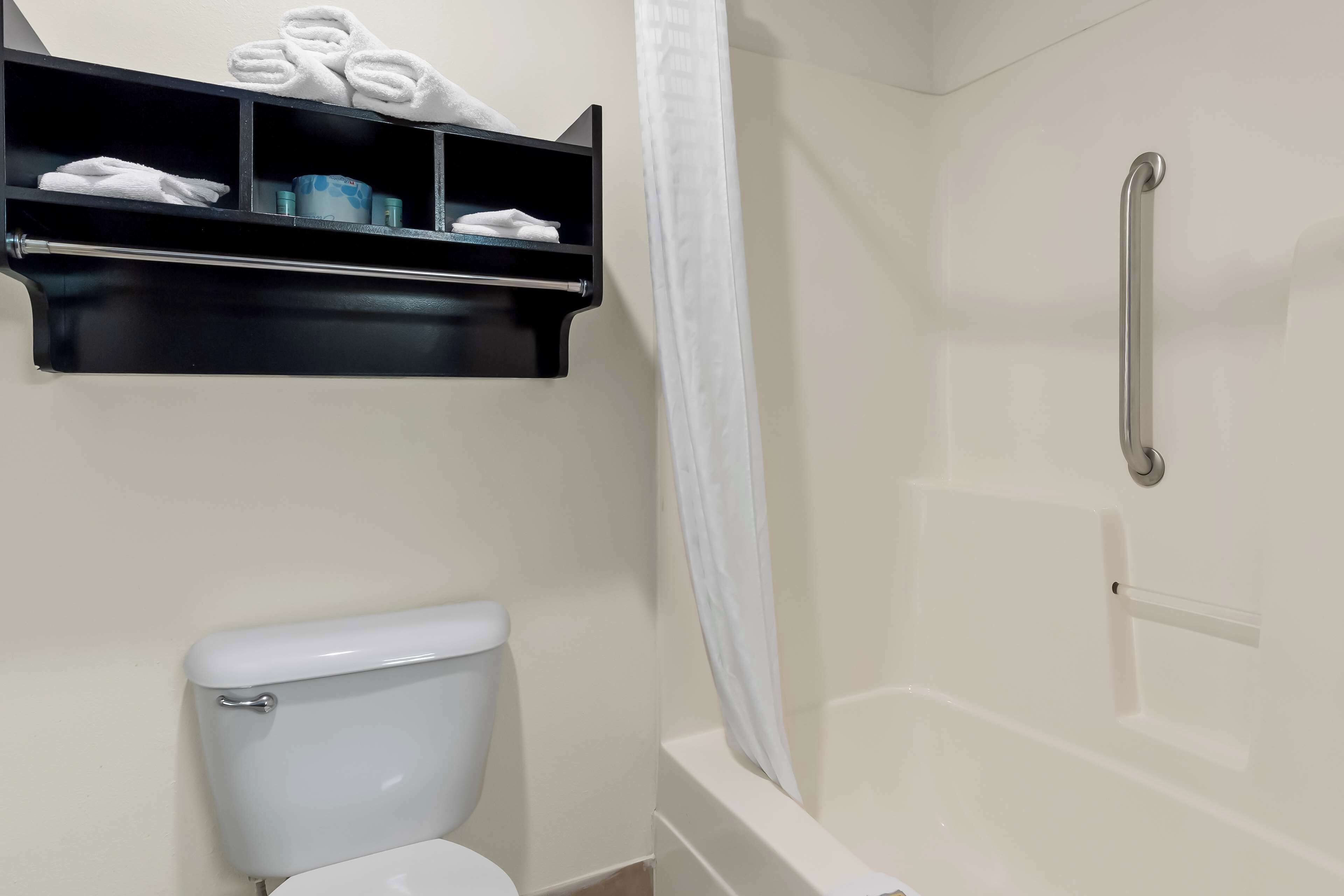 Un baño con inodoro, ducha con barra de apoyo y estante con toallas y artículos de aseo.