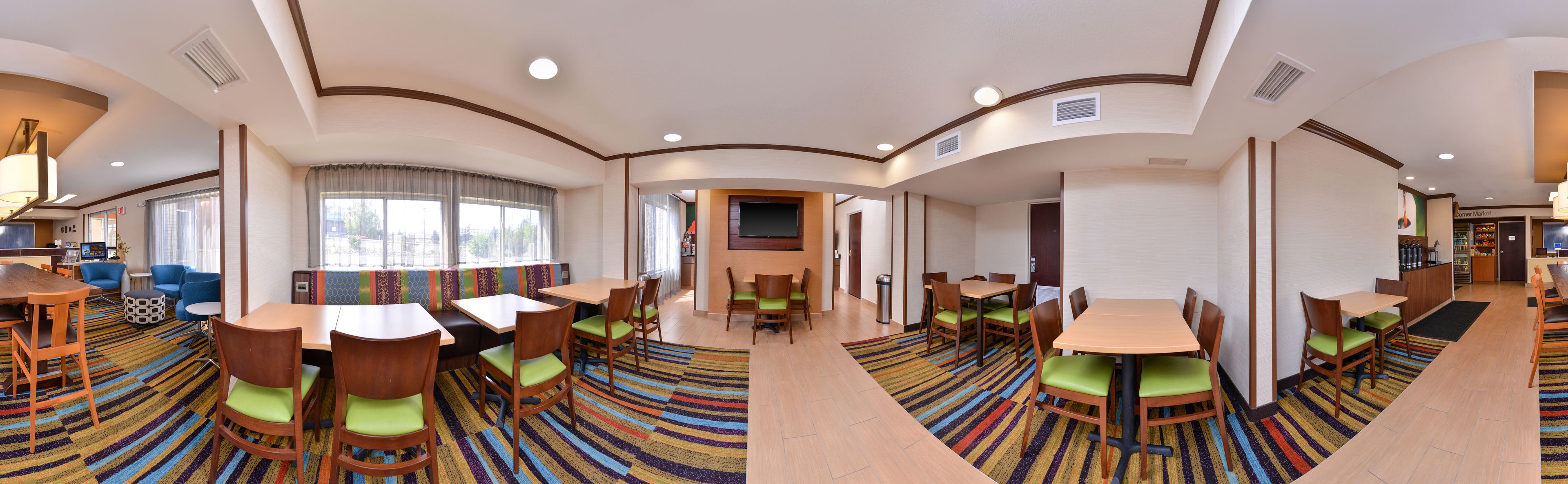 A hotel dining area with multiple tables and chairs, a TV mounted on the wall, and a large window with curtains.