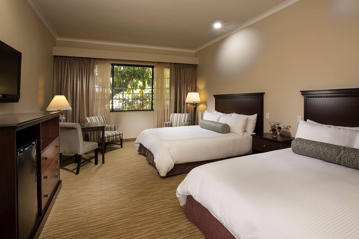 A hotel room with two beds, a desk, a chair, a TV, and a window with curtains.