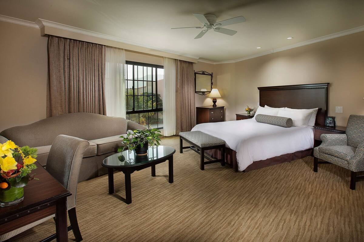A hotel room with a bed, a sofa, a dining table with a vase of flowers, a small table with a lamp, and a chair.