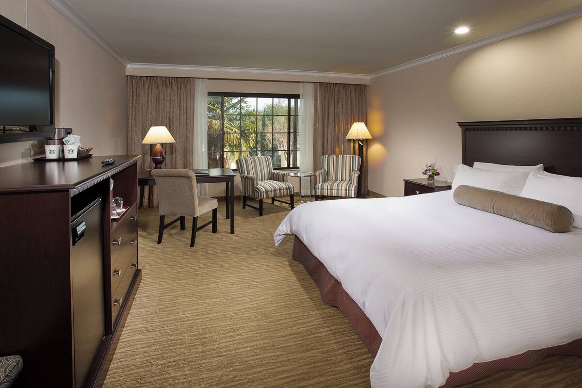 A hotel room with a large bed, a desk with two chairs, a television, and a window with curtains.