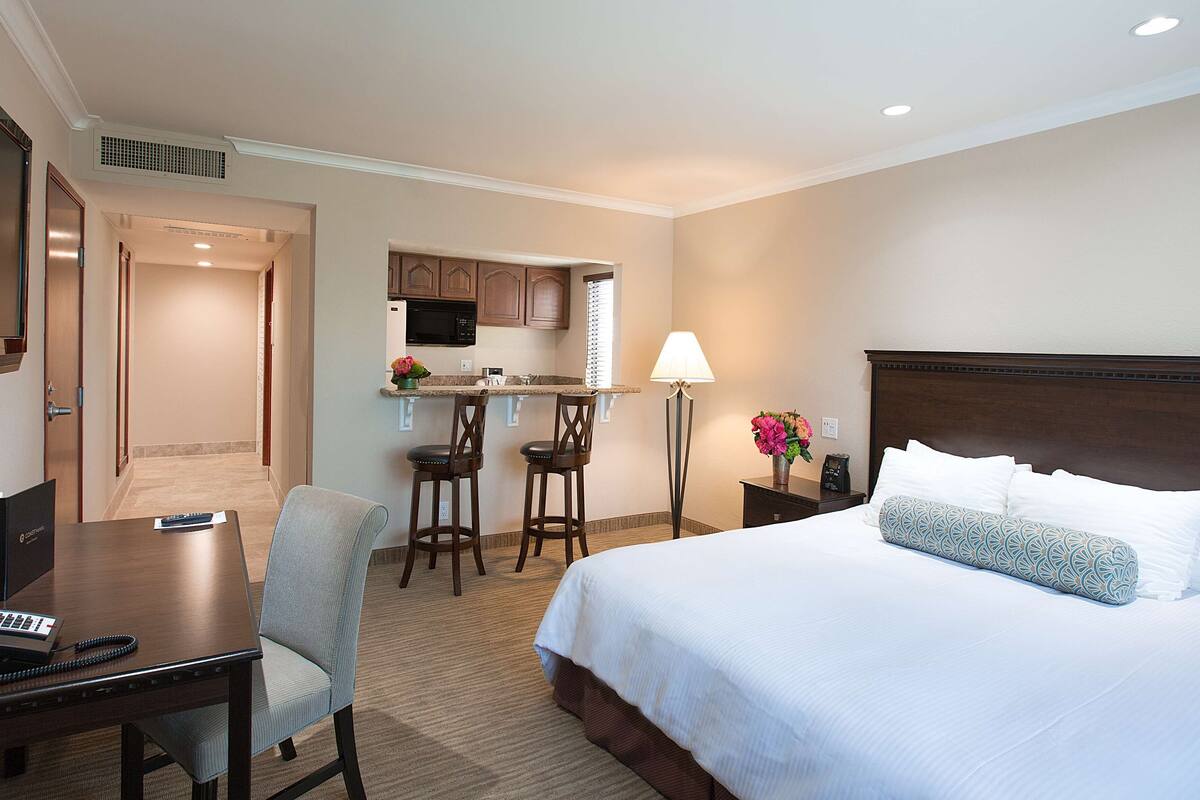 A hotel room with a bed, desk, chairs, a small kitchenette, and a nightstand with a lamp.
