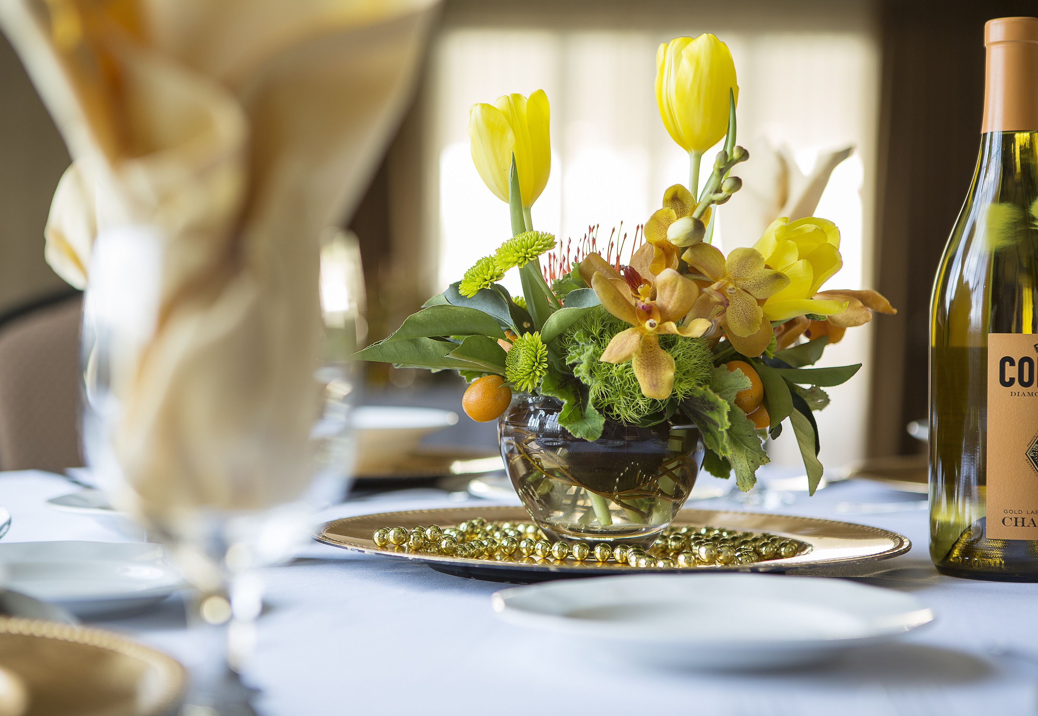 A table set with a glass vase of flowers, a bottle of wine, and gold-plated dinnerware.