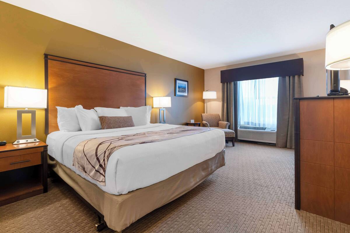 A hotel room with a large bed, two bedside tables with lamps, a chair, a desk, and a large window with curtains.
