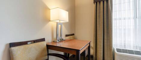 A hotel room with a wooden table, two chairs, a lamp, and an air conditioning unit near a window with curtains.