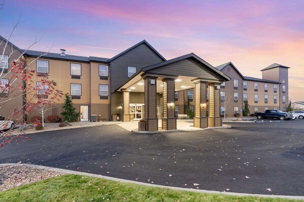 A multi-story hotel with a central entrance, parking lot, and a clear sky.