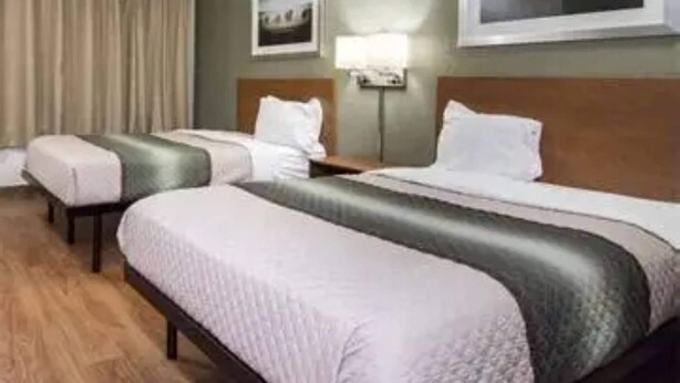 A hotel room with two beds, a nightstand, a lamp, and a framed picture on the wall. - Suburban Studios Durham-Raleigh (Durham)