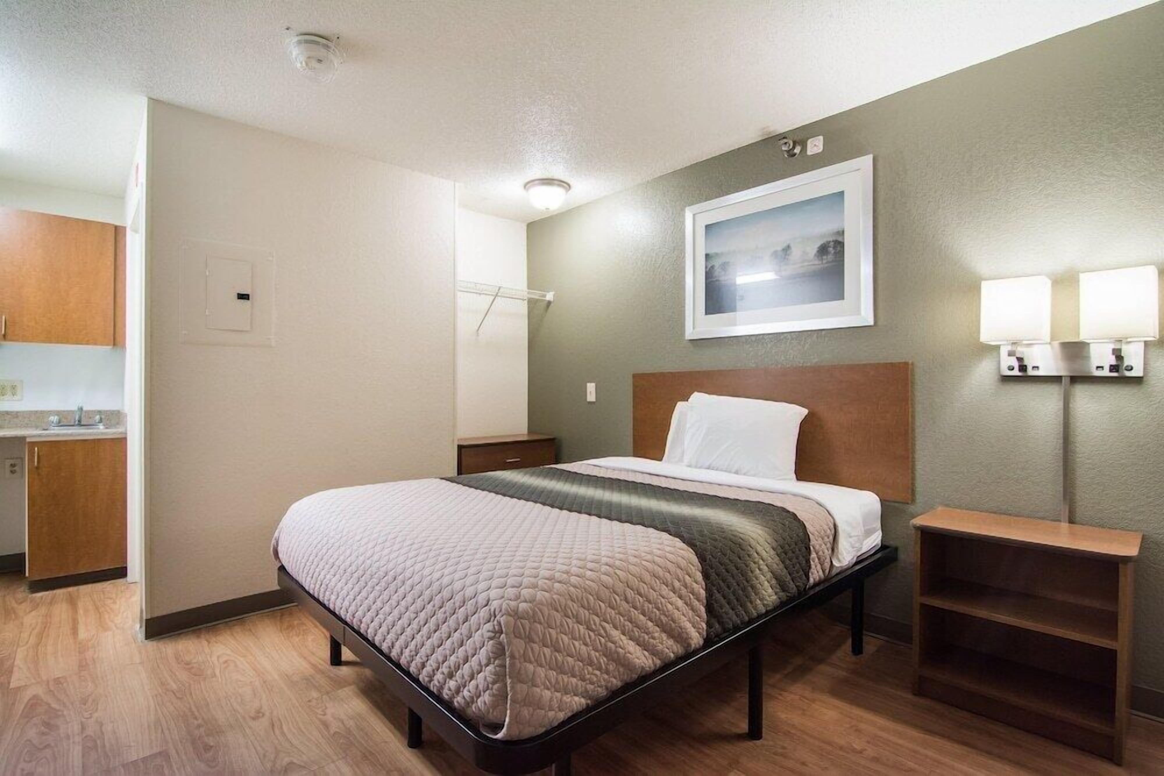A hotel room with a bed, a nightstand, a wall-mounted lamp, and a painting above the bed.