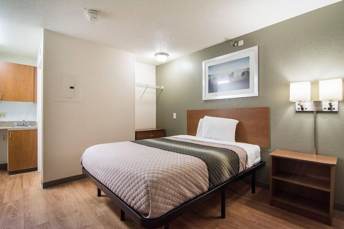 A hotel room with a bed, a nightstand, a wall-mounted lamp, and a painting above the bed.