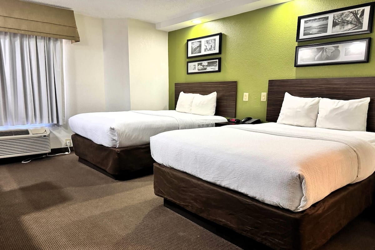 A hotel room with two beds, a large window with curtains, a wall-mounted air conditioner, and framed pictures on the wall.