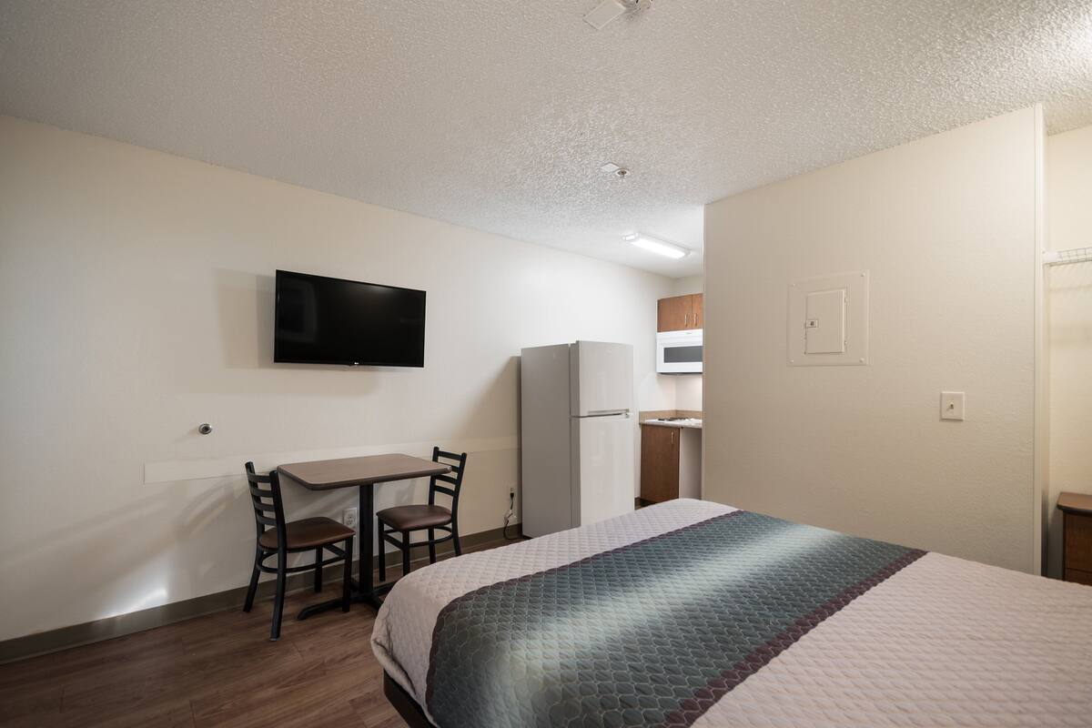 A hotel room with a bed, a desk with a chair, a television, and a kitchenette.