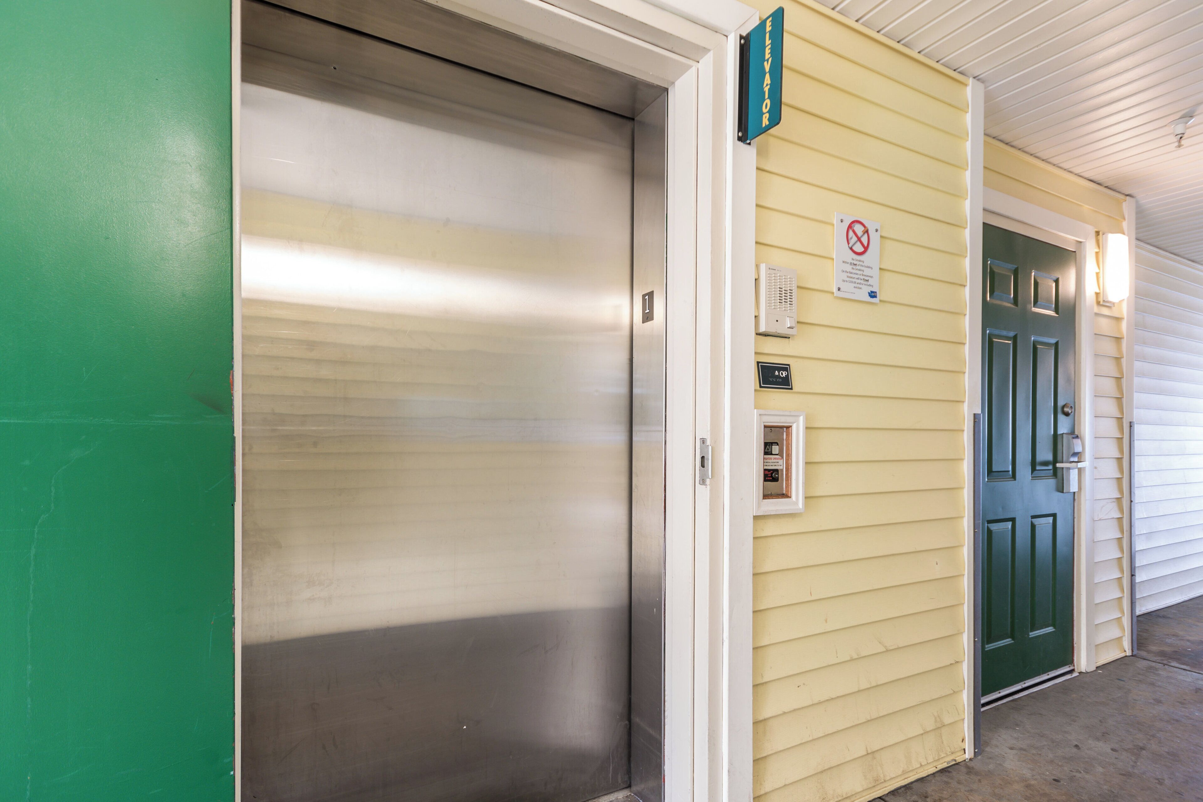 A closed elevator door with a green sign above it, a doorbell, and a smoke detector on the wall.
