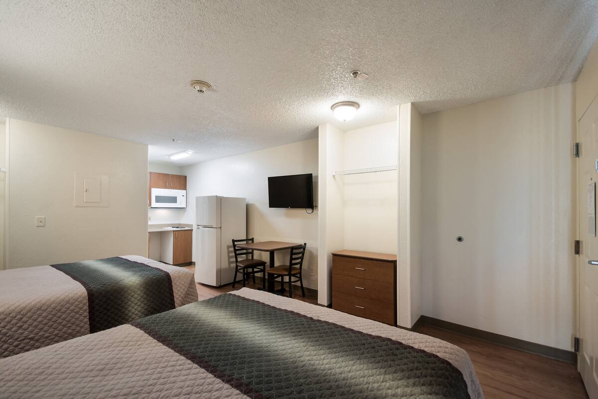 A hotel room with two beds, a desk, a chair, a television, and a kitchenette.