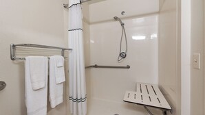 Zona de ducha con mampara de vidrio, cabezal de ducha manual, toallero con toallas dobladas y barra de apoyo.