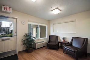 A reception area with an exit sign, a clock, a window, a potted plant, two leather chairs, and a small table. - HomeTowne Studios By Red Roof Seattle - Kent/ Des Moines (Kent)