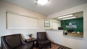 Un área de recepción con dos sillones de cuero, una mesita con una planta, un mostrador de recepción con una computadora y una caja registradora, y una pared verde con un letrero que dice 'HomeTown Studios'.