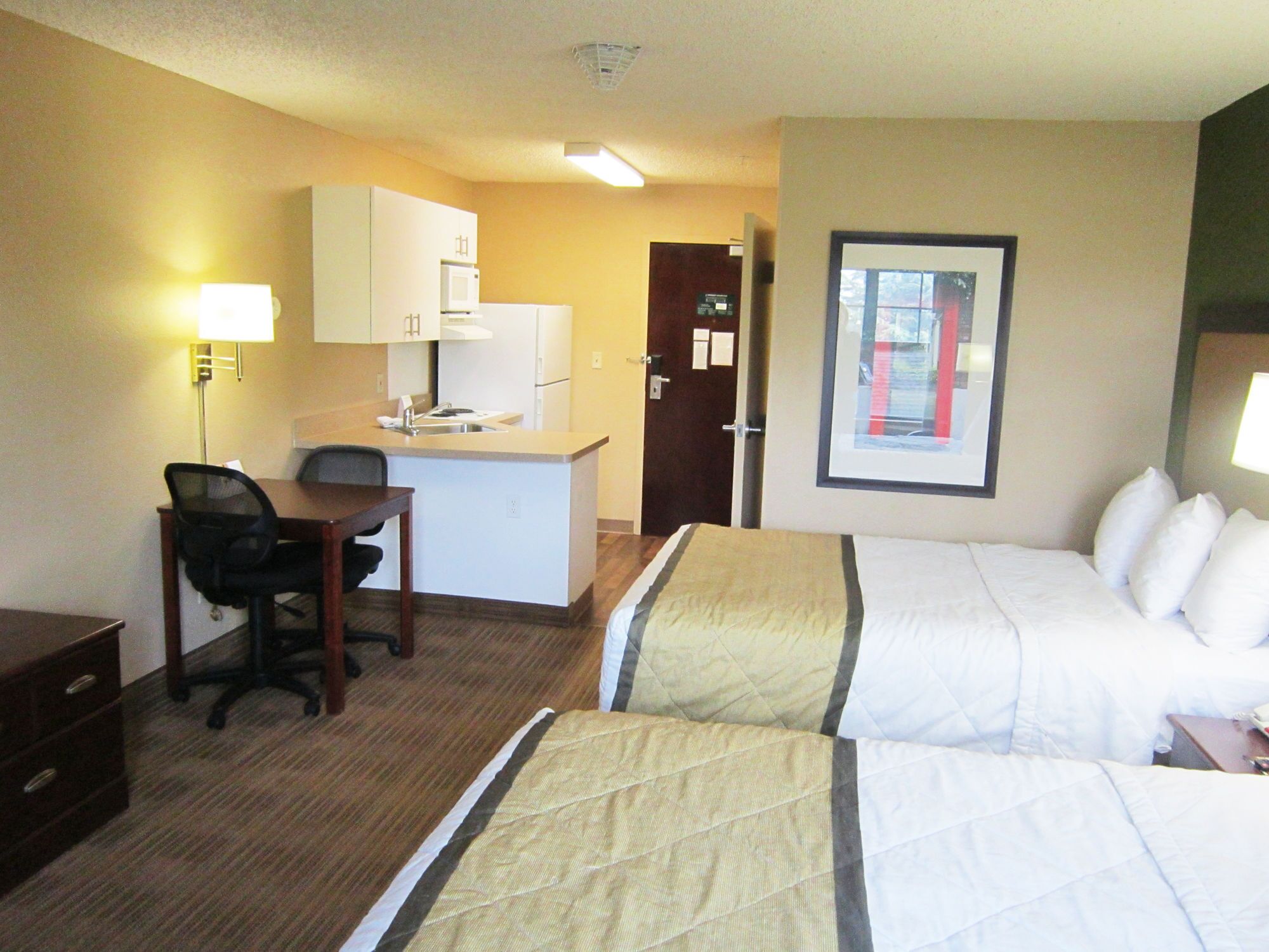 A hotel room with two beds, a desk, a chair, a kitchenette, and a framed picture on the wall.