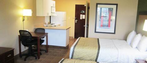 A hotel room with two beds, a desk, a chair, a kitchenette, and a framed picture on the wall.