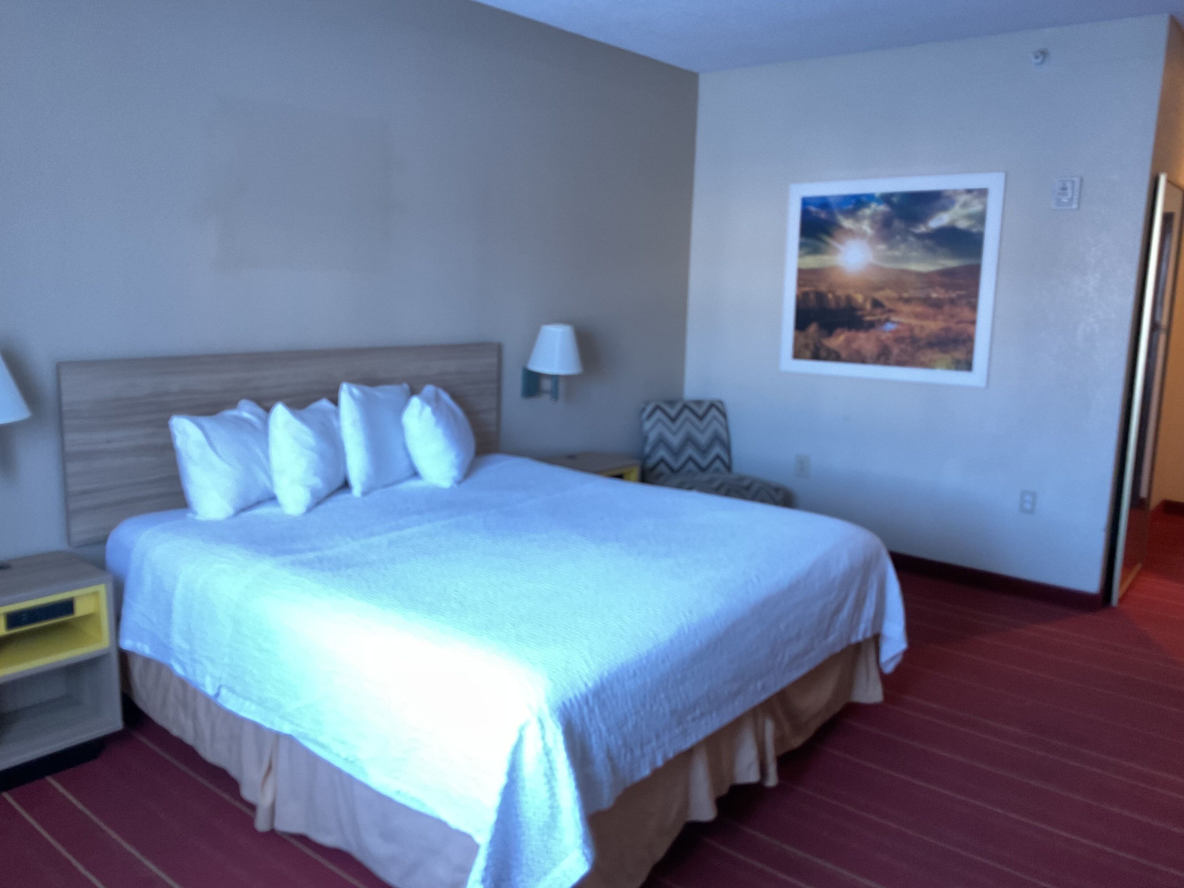 Una camera d'albergo con un letto, un comodino, una lampada, un quadro appeso e un pavimento coperto da un tappeto rosso.