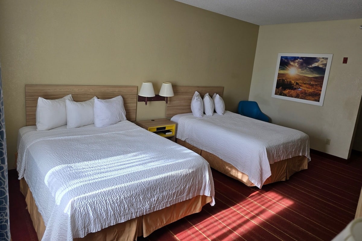 A hotel room with two beds, a nightstand with lamps, a chair, and a painting on the wall.