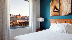 A hotel room with a large bed, a nightstand, a lamp, and a view of a cityscape through a window.