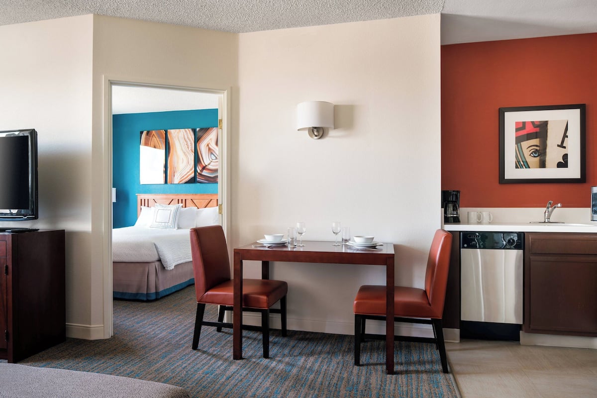 Een hotelkamer met een bed, een televisie, een eettafel met stoelen, een keukenhoek en een schilderij aan de muur.