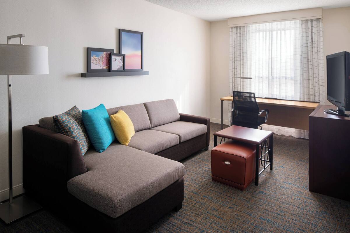 Een moderne hotelkamer met een hoekbank, een klein bureau met een computer, een flatscreen televisie en een lamp.