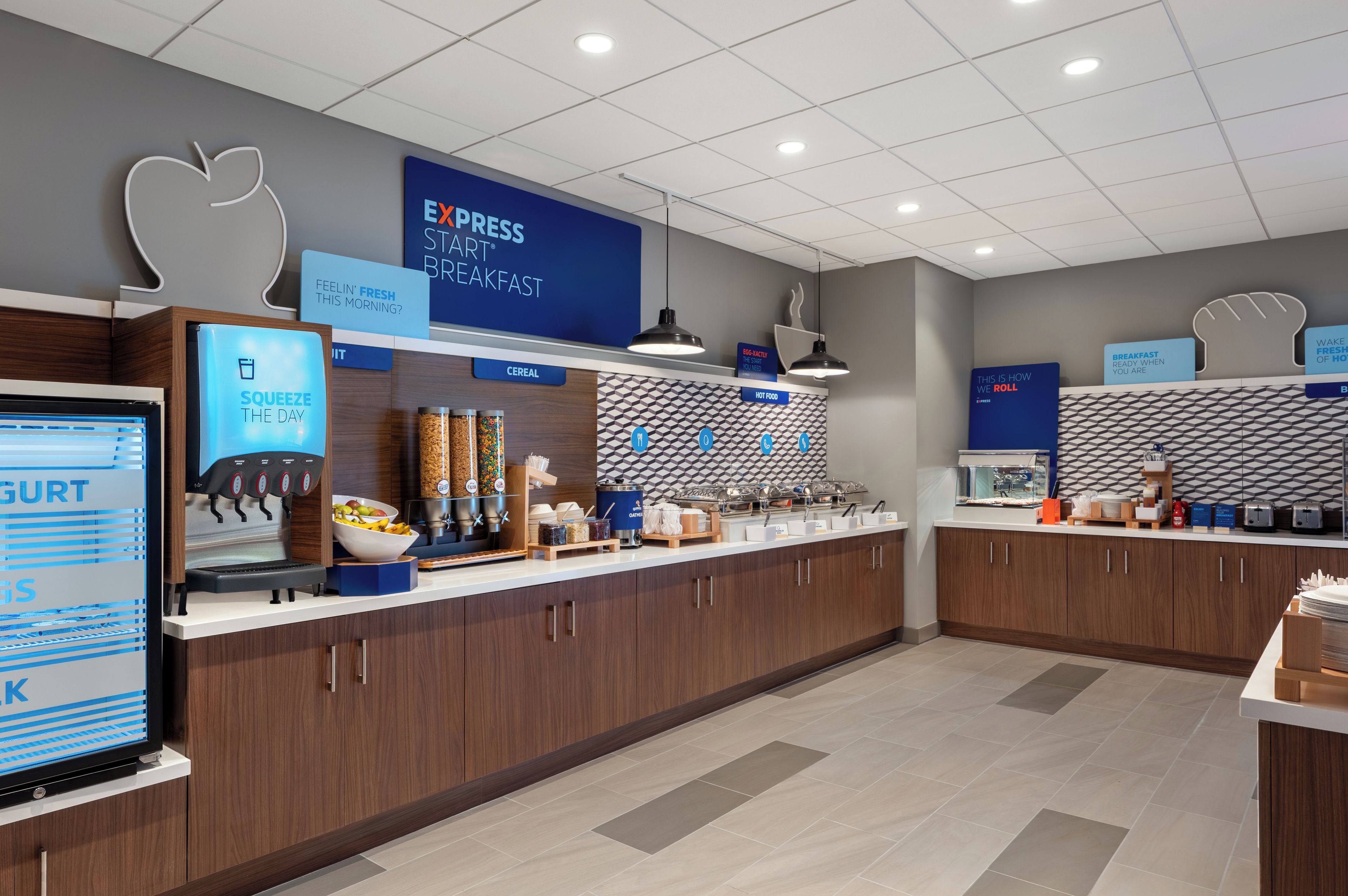 A breakfast area with a digital screen, self-serve beverage station, and various food options.
