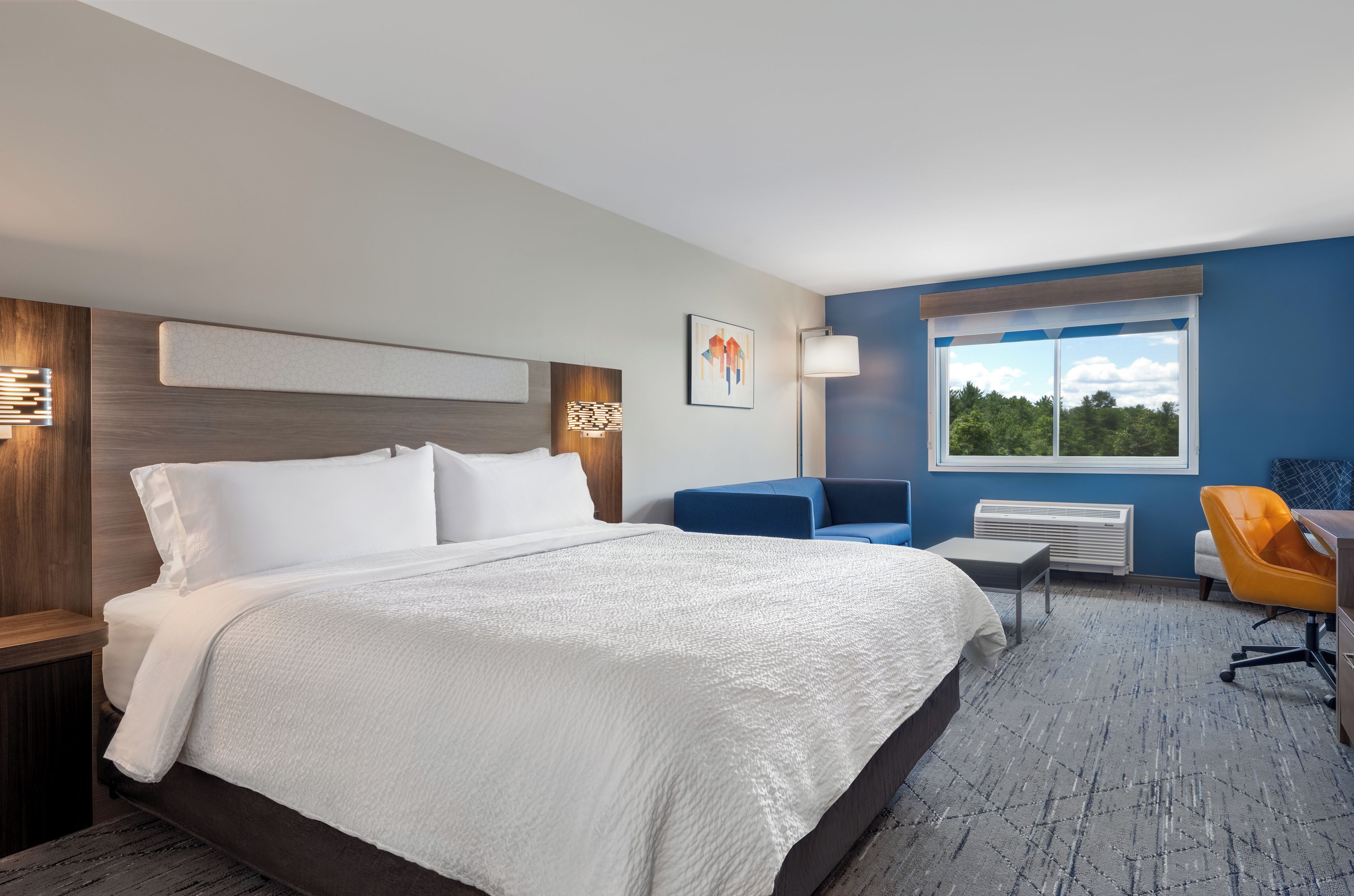 Quarto de hotel moderno com uma cama grande, uma escrivaninha, uma cadeira e uma janela com vista para árvores.