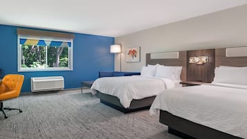 A hotel room with two beds, a blue accent wall, a window with a view of trees, a chair, and a heater.