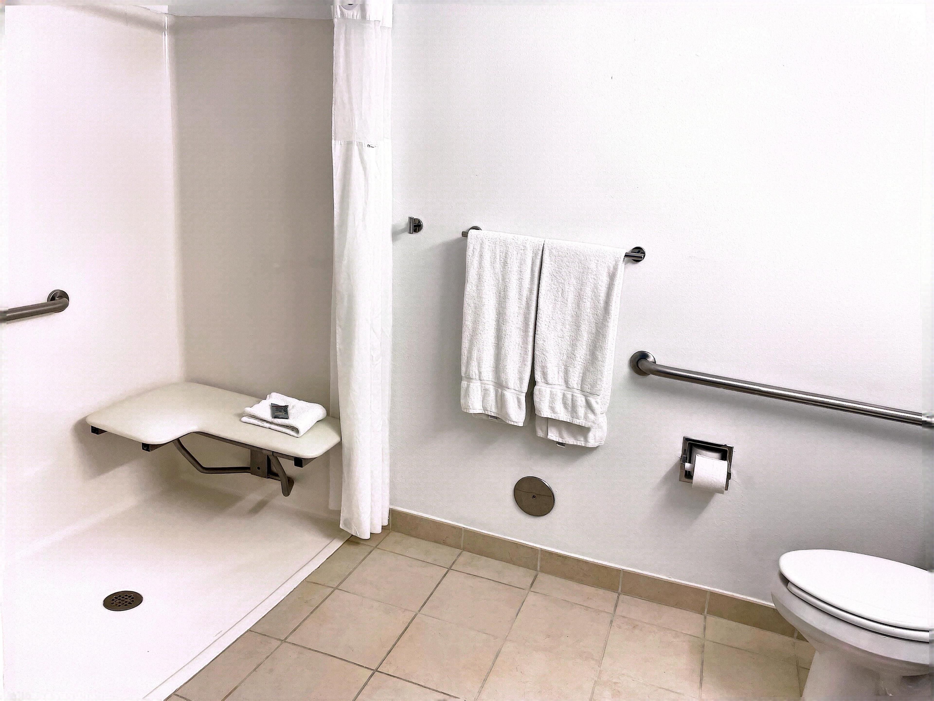 A bathroom with a shower, a toilet, a grab bar, a towel rack, and a small bench.