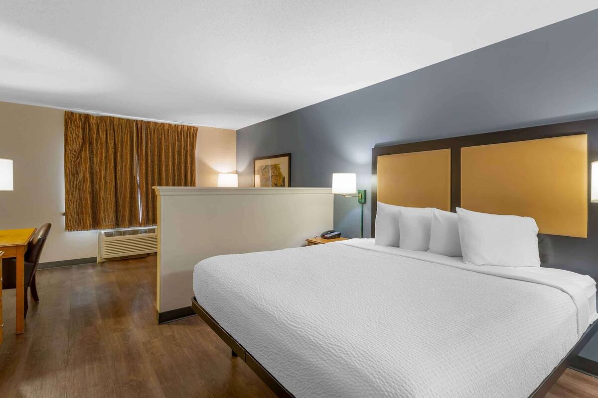 Een hotelkamer met een groot bed, een bureau en een stoel. Er is een raam met gordijnen, een wandlamp en een schilderij aan de muur.
