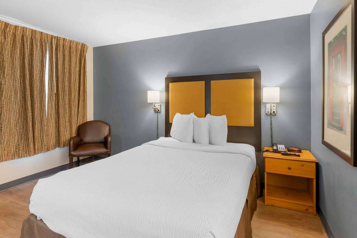 Een hotelkamer met een groot bed, twee bedlampen, een houten nachtkastje, een bruine leren fauteuil en een gordijn met dessin.