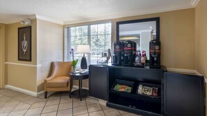 Habitación de hotel con pared color crema, un ventanal con persianas, una estación de café negro, un sillón de cuero y una mesita con una lámpara.