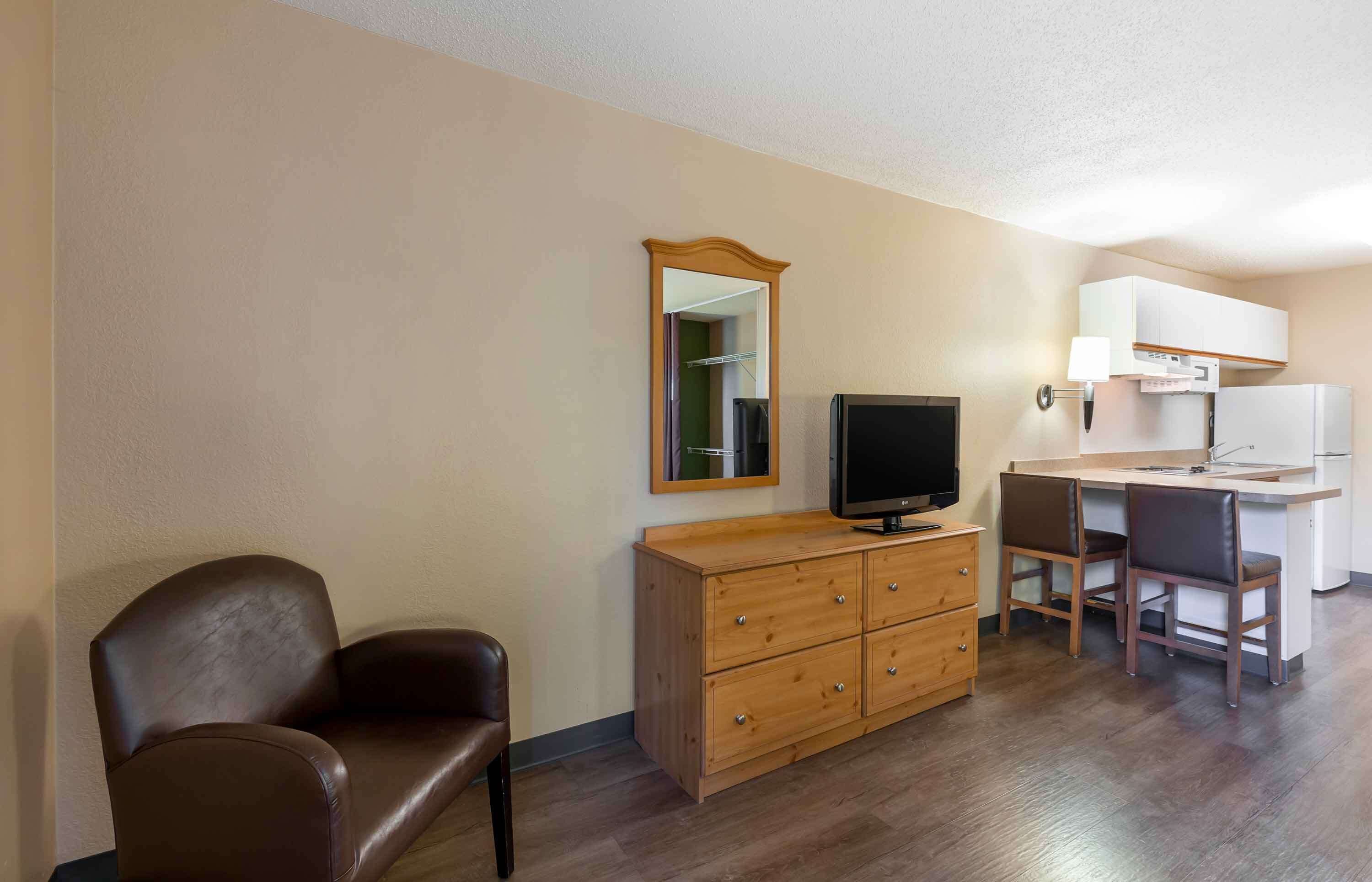 A compact hotel room with a single brown leather chair, a wooden dresser with a flat-screen TV, a small kitchenette, and a mirror on the wall.